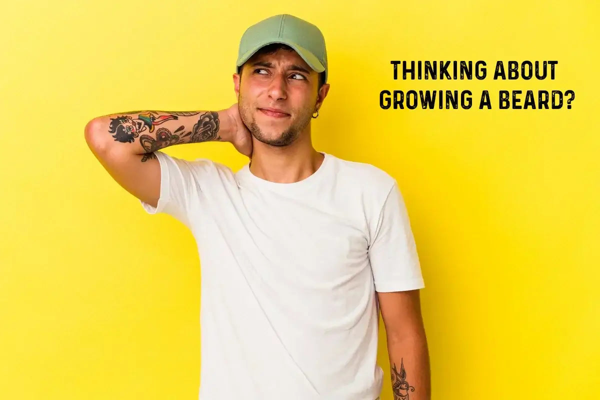 Young man with tattoos wearing a white t-shirt and green cap, looking uncertain while scratching his neck, standing in front of a bright yellow background with the text “Thinking about growing a beard?” next to him.