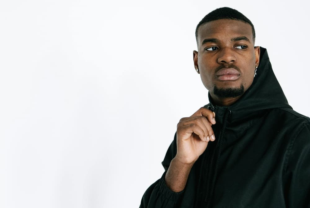 A stylish man wearing a black hoodie, posing thoughtfully against a plain white background.