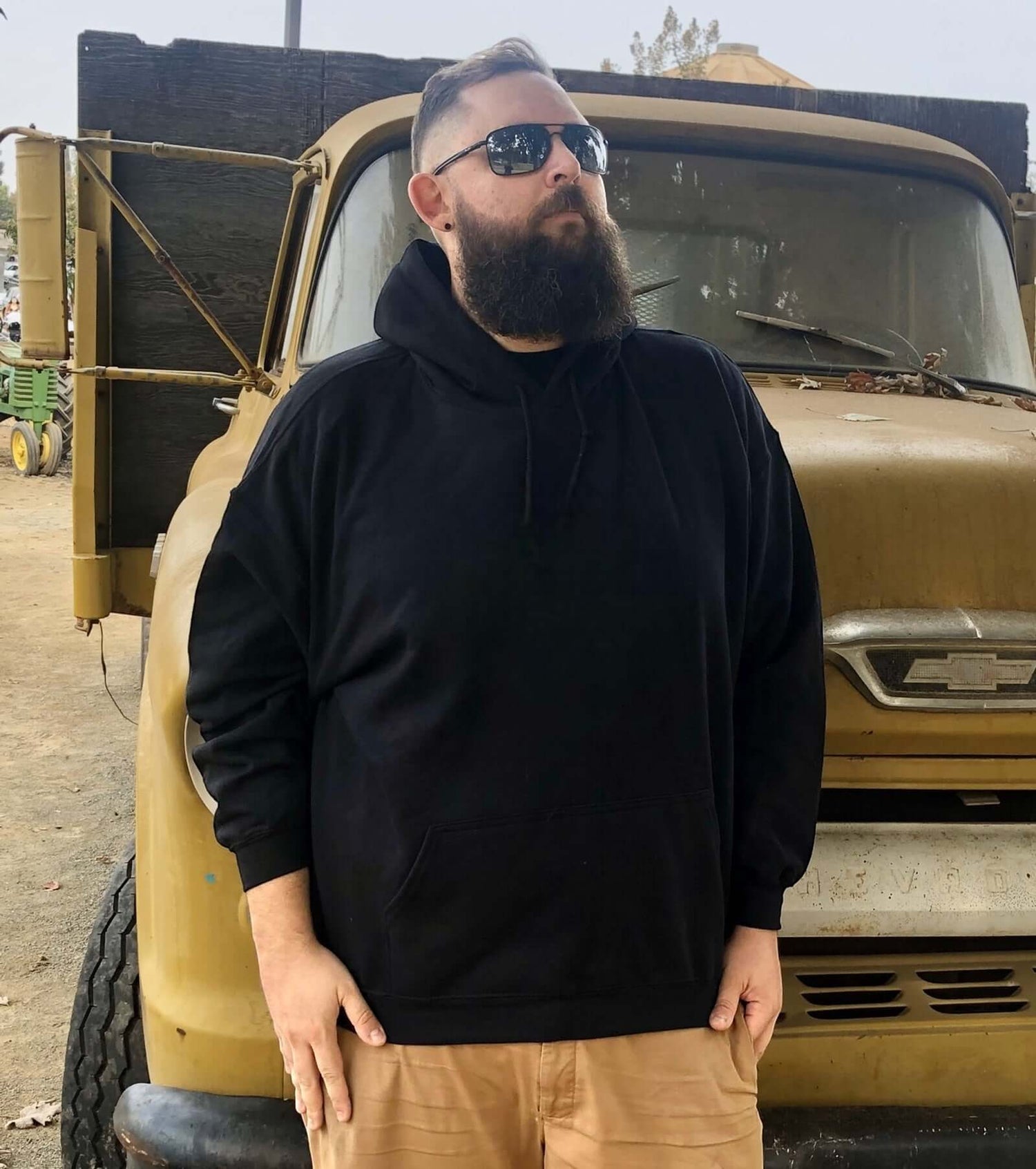 Man wearing a black hoodie stands confidently in front of an old truck, showcasing his beard.