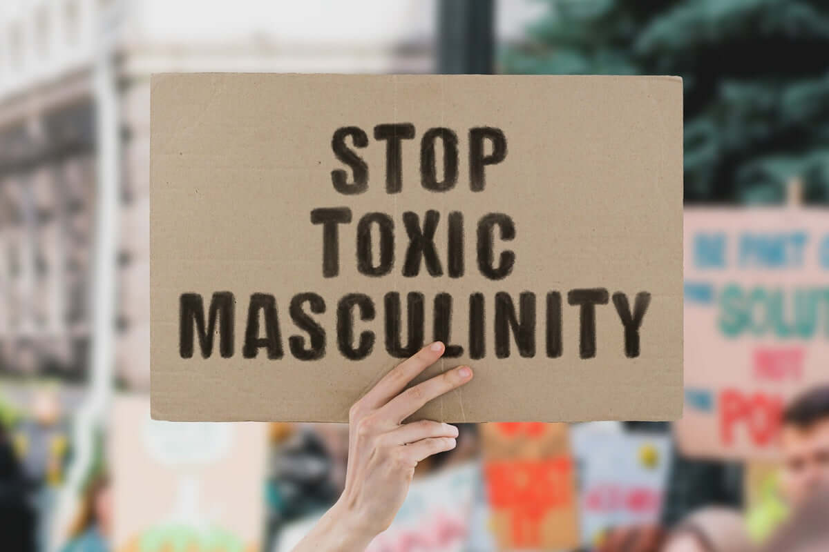 Hand holding a cardboard sign that reads 'STOP TOXIC MASCULINITY' in bold black stencil letters, with a blurred protest scene in the background.