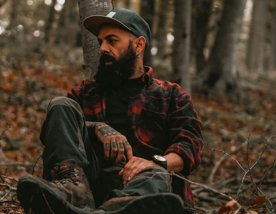 Man with a full beard wearing a flannel shirt and cap, sitting in a forest, embodying rebel grooming style.
