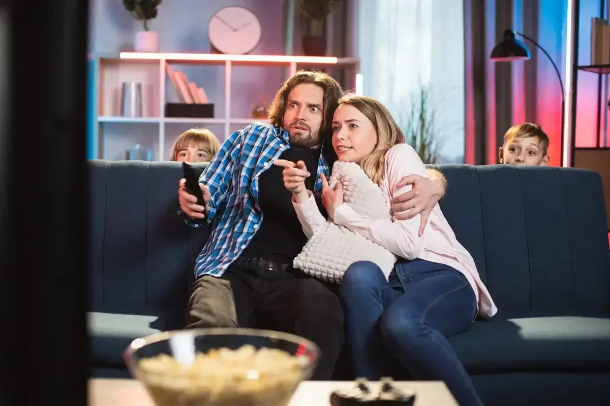 Family watching a scary movie on the couch, parents hugging tightly with shocked expressions