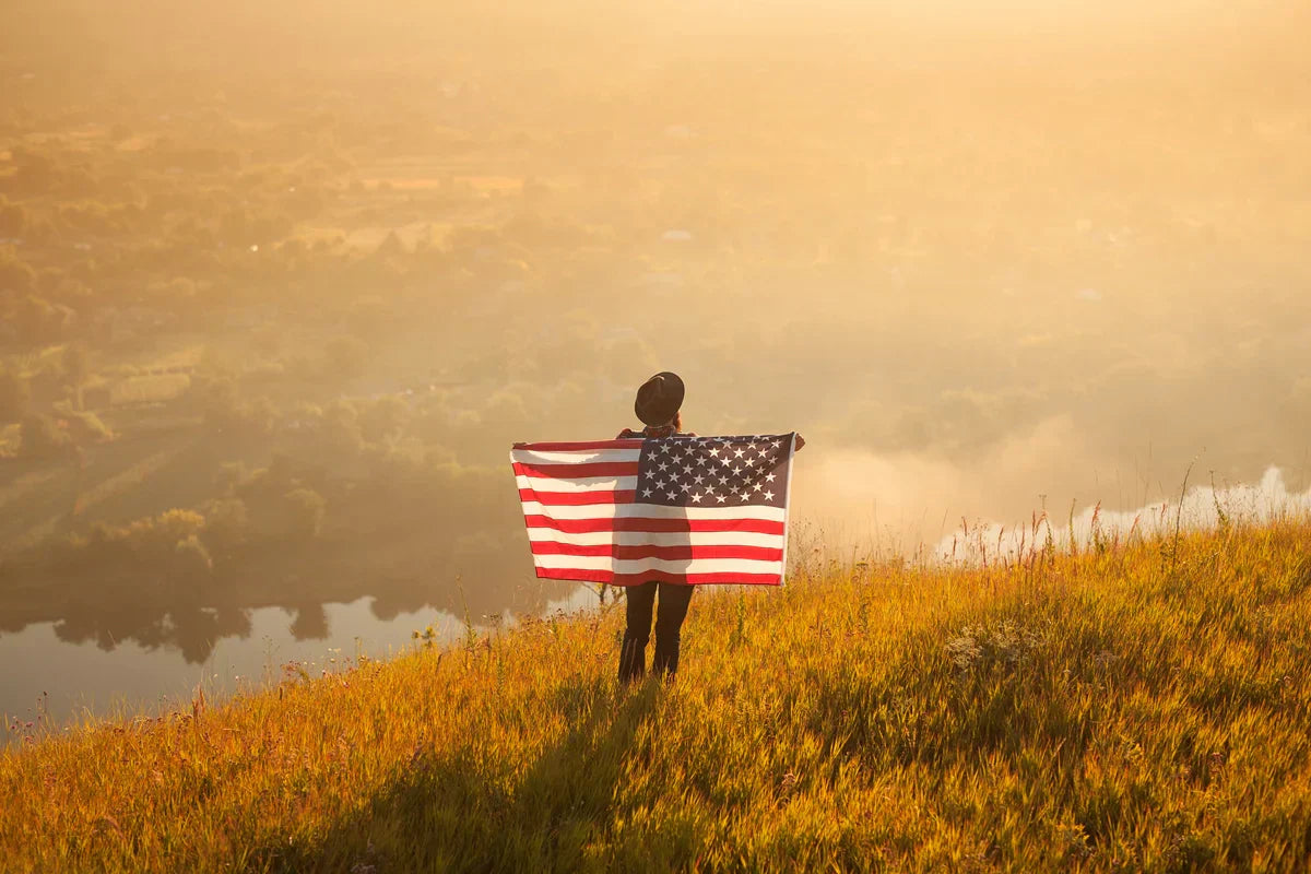 Person seen from behind holding an American flag open across their back, standing on a golden hillside overlooking a misty river valley at sunset