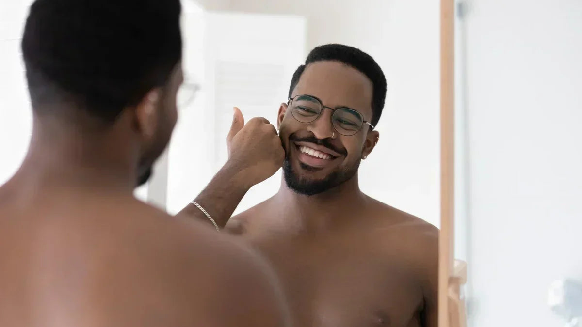 Smiling man with short beard admiring his reflection while gently touching his cheek in bathroom mirror.