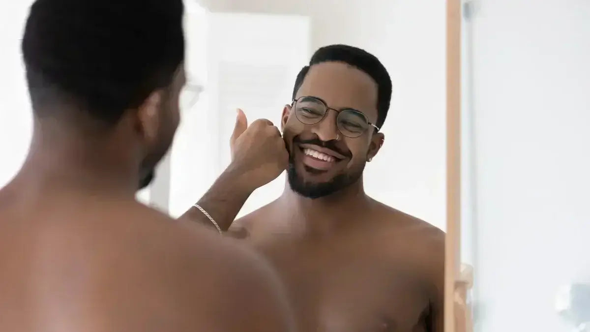 Smiling man with short beard admiring his reflection while gently touching his cheek in bathroom mirror.