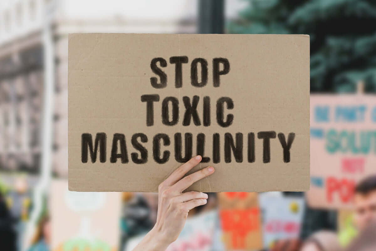 Hand holding a cardboard sign that reads 'STOP TOXIC MASCULINITY' in bold black stencil letters, with a blurred protest scene in the background.