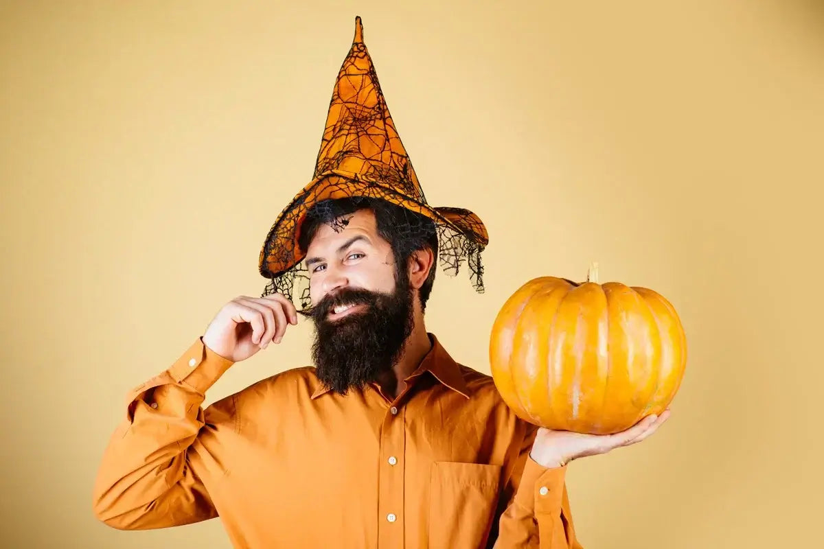Man with styled beard in Halloween witch hat holding a pumpkin against a beige background.