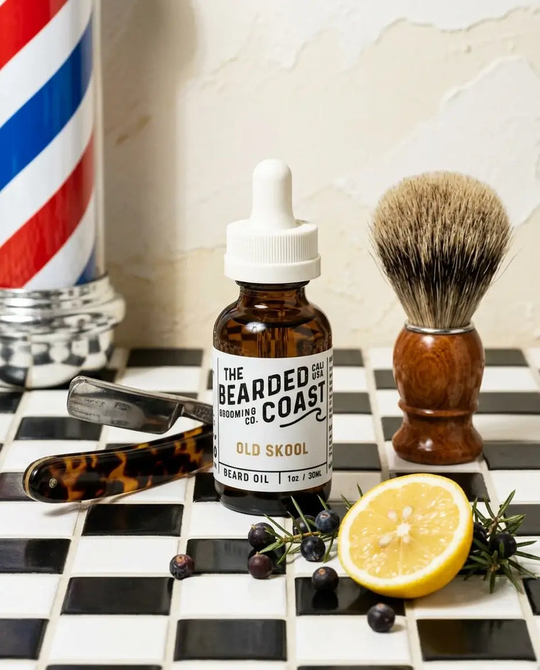Amber dropper bottle of Bearded Coast Grooming Co. Old Skool Beard Oil, 1oz/30ml, styled on a classic black and white checkered tile surface alongside a straight razor, shaving brush, fresh lemon, juniper berries, and rosemary sprigs, with a vintage barber pole in the background.