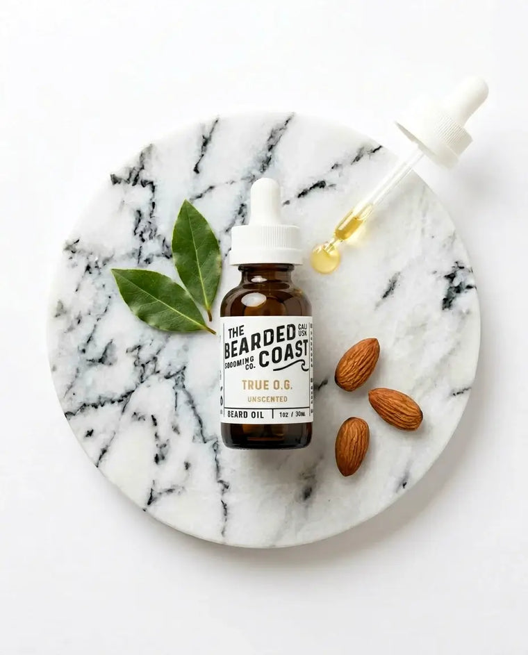 Overhead flat lay of Bearded Coast Grooming Co. True O.G. Unscented Beard Oil, 1oz/30ml, on a round white marble slab surrounded by fresh bay leaves, raw almonds, and a glass dropper with a drop of golden oil.