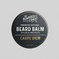 Top-down view of The Bearded Coast Grooming Co. Carpe Diem Premium Natural Beard Balm — a 2 oz / 57g dark charcoal metal tin with gold and white typography on a gray background