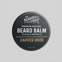 Top-down view of The Bearded Coast Grooming Co. Dapper Dude Premium Natural Beard Balm — a 2 oz / 57g dark charcoal metal tin with gold and white typography on a gray background