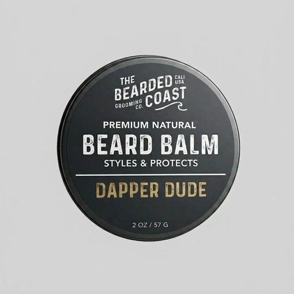 Top-down view of The Bearded Coast Grooming Co. Dapper Dude Premium Natural Beard Balm — a 2 oz / 57g dark charcoal metal tin with gold and white typography on a gray background