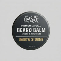 Top-down view of The Bearded Coast Grooming Co. Dark'n Stormy Premium Natural Beard Balm — a 2 oz / 57g dark charcoal metal tin with gold and white typography on a gray background