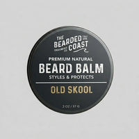 Top-down view of The Bearded Coast Grooming Co. Old Skool Premium Natural Beard Balm — a 2 oz / 57g dark charcoal metal tin with gold and white typography on a gray background
