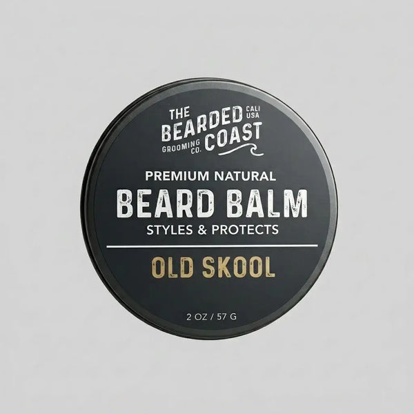 Top-down view of The Bearded Coast Grooming Co. Old Skool Premium Natural Beard Balm — a 2 oz / 57g dark charcoal metal tin with gold and white typography on a gray background