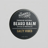 Top-down view of The Bearded Coast Grooming Co. Salty Vibes Premium Natural Beard Balm — a 2 oz / 57g dark charcoal metal tin with gold and white typography on a gray background
