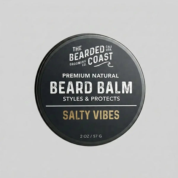Top-down view of The Bearded Coast Grooming Co. Salty Vibes Premium Natural Beard Balm — a 2 oz / 57g dark charcoal metal tin with gold and white typography on a gray background