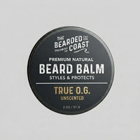 Top-down view of The Bearded Coast Grooming Co. True O.G. Premium Natural Beard Balm — a 2 oz / 57g dark charcoal metal tin with gold and white typography on a gray background