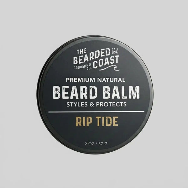 Top-down view of The Bearded Coast Grooming Co. Rip Tide Premium Natural Beard Balm — a 2 oz / 57g dark charcoal metal tin with gold and white typography on a gray background